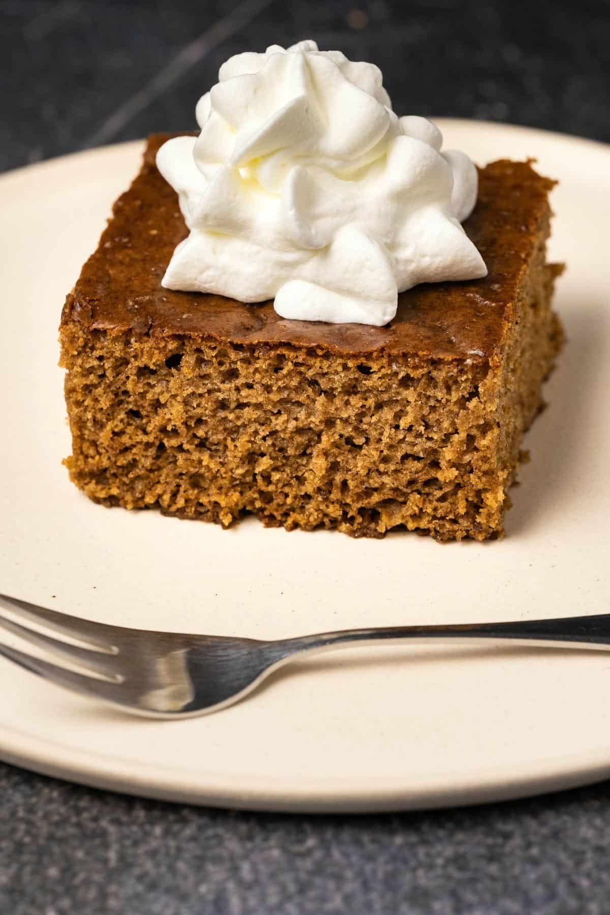 Slice of gingerbread cake with whipped cream, on a white plate with a cake fork. 