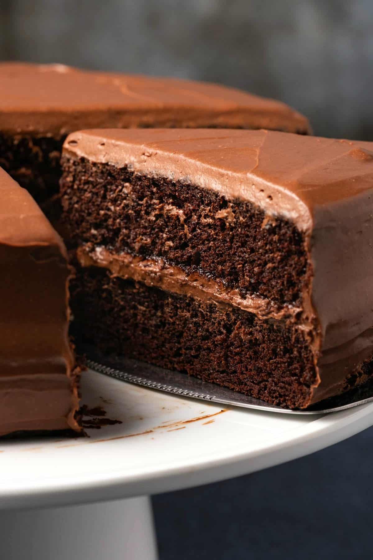 Chocolate cake on a white cake stand with one slice cut and ready to serve. 