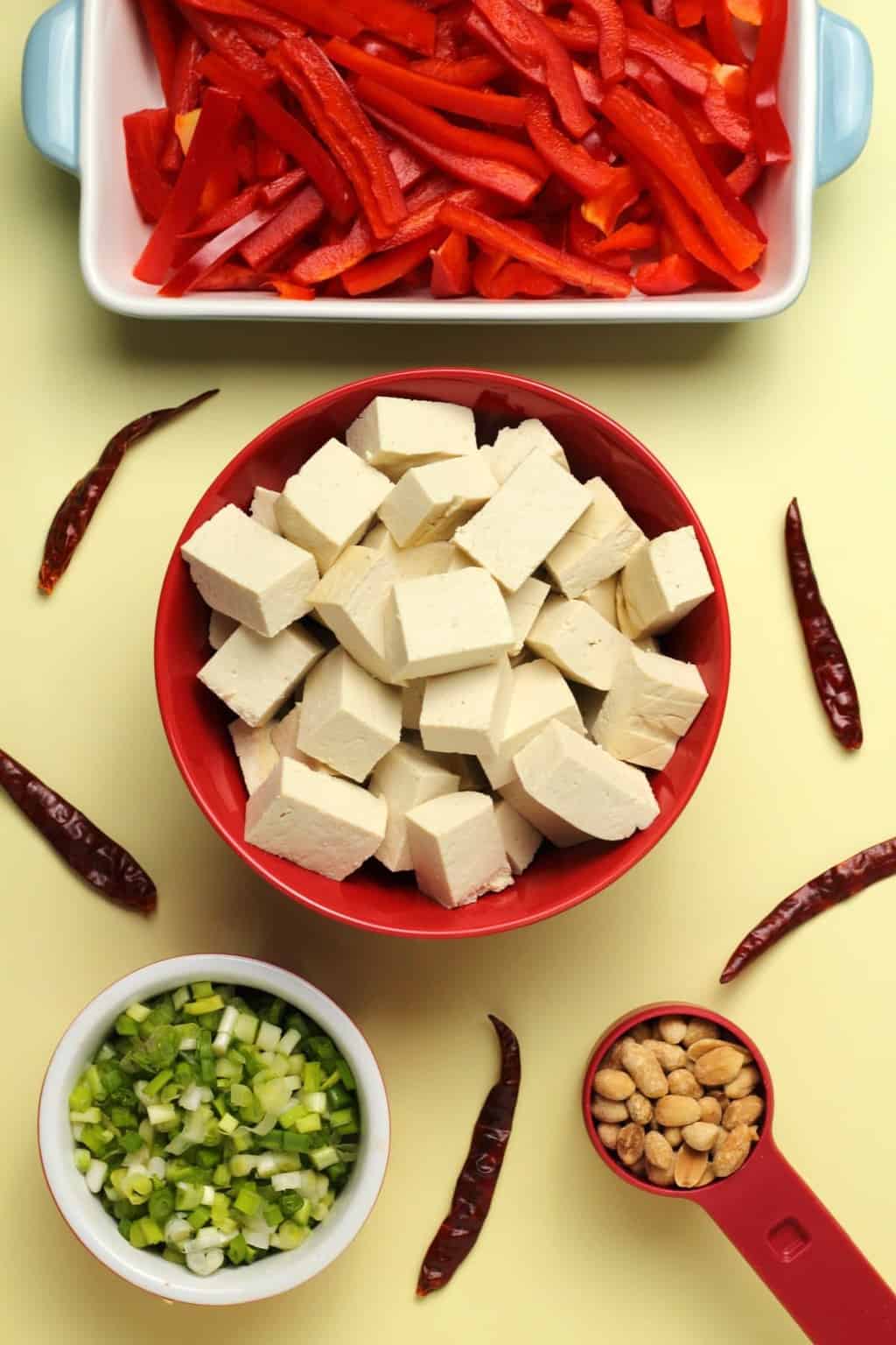 Ingredients for kung pao tofu, cubes of tofu in a bowl, sliced red peppers, dried chilies, chopped spring onion and roasted salted peanuts.
