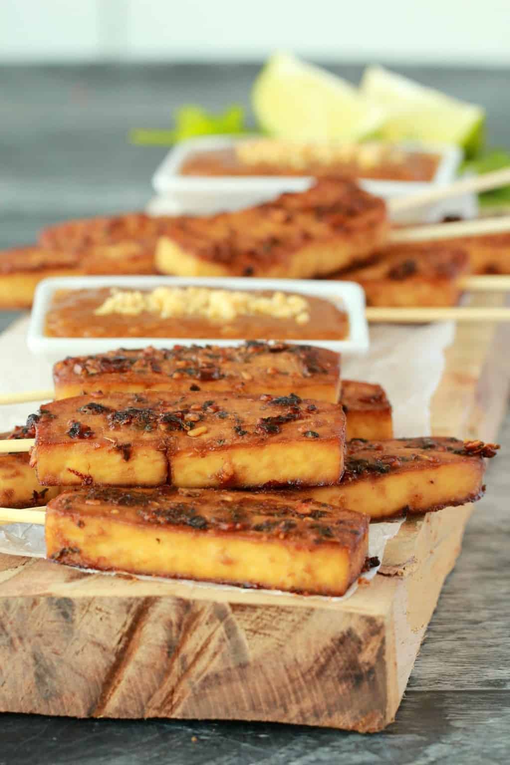 Baked tofu satay on a wooden board with peanut sauce in a white dish. 