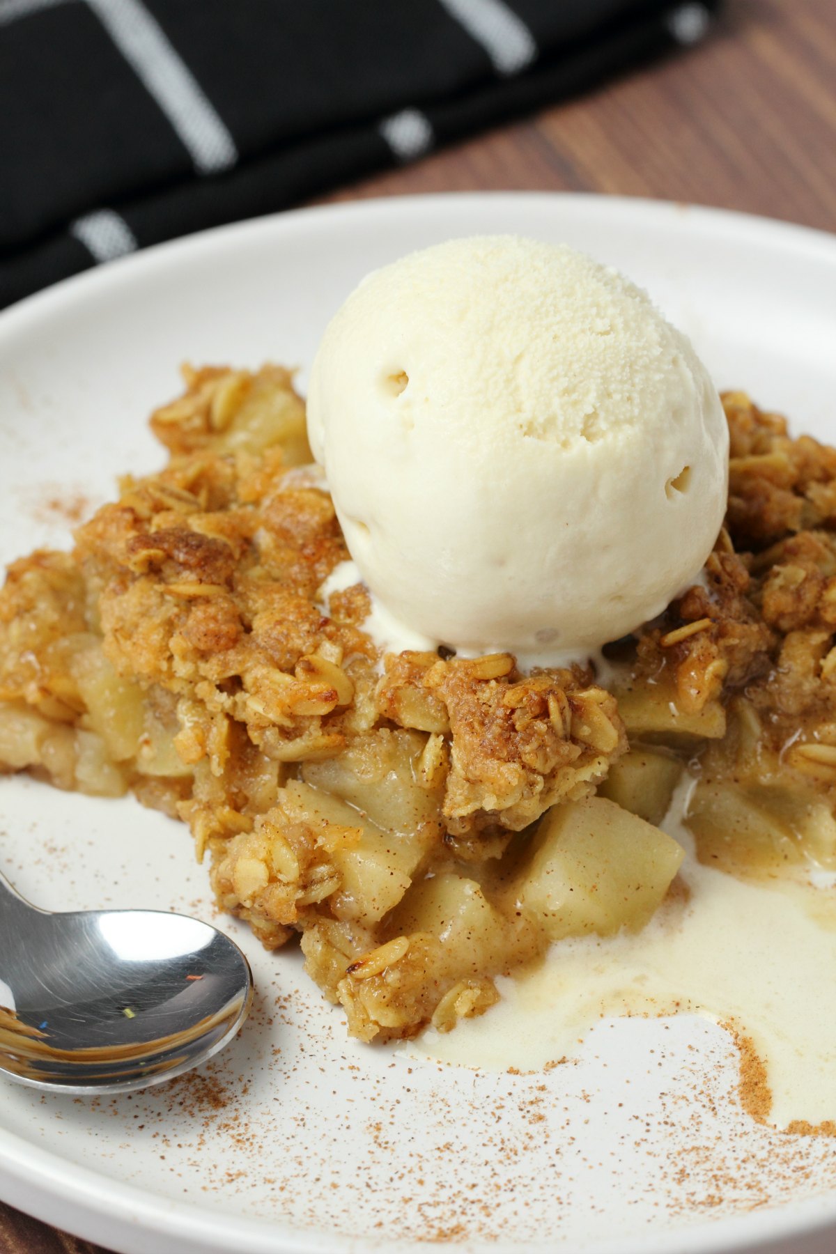 A slice of vegan apple crisp with a scoop of vegan vanilla ice cream on top on a white plate. 