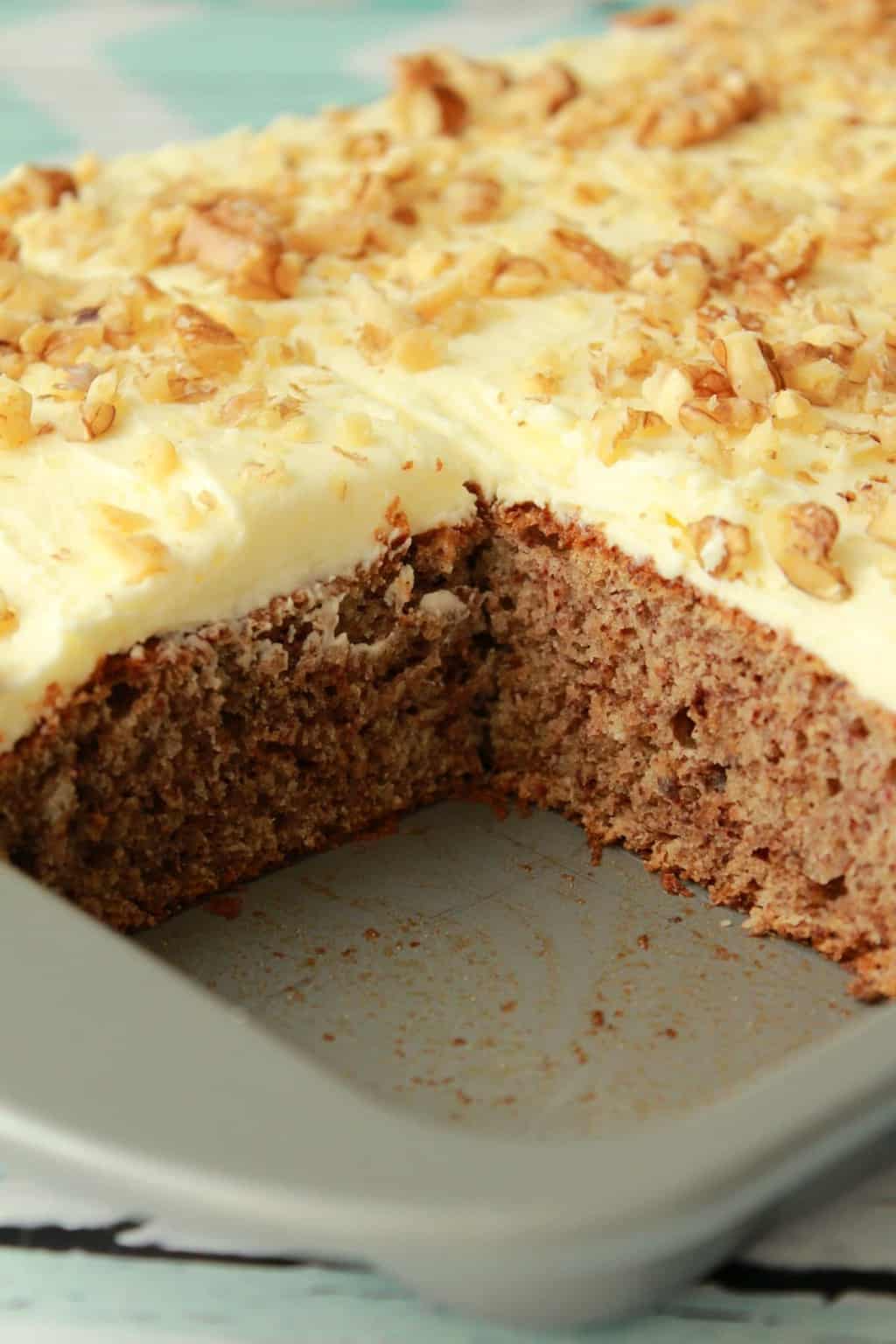 Vegan banana cake topped with lemon frosting and crushed walnuts in a quarter sheet pan with one slice removed. 
