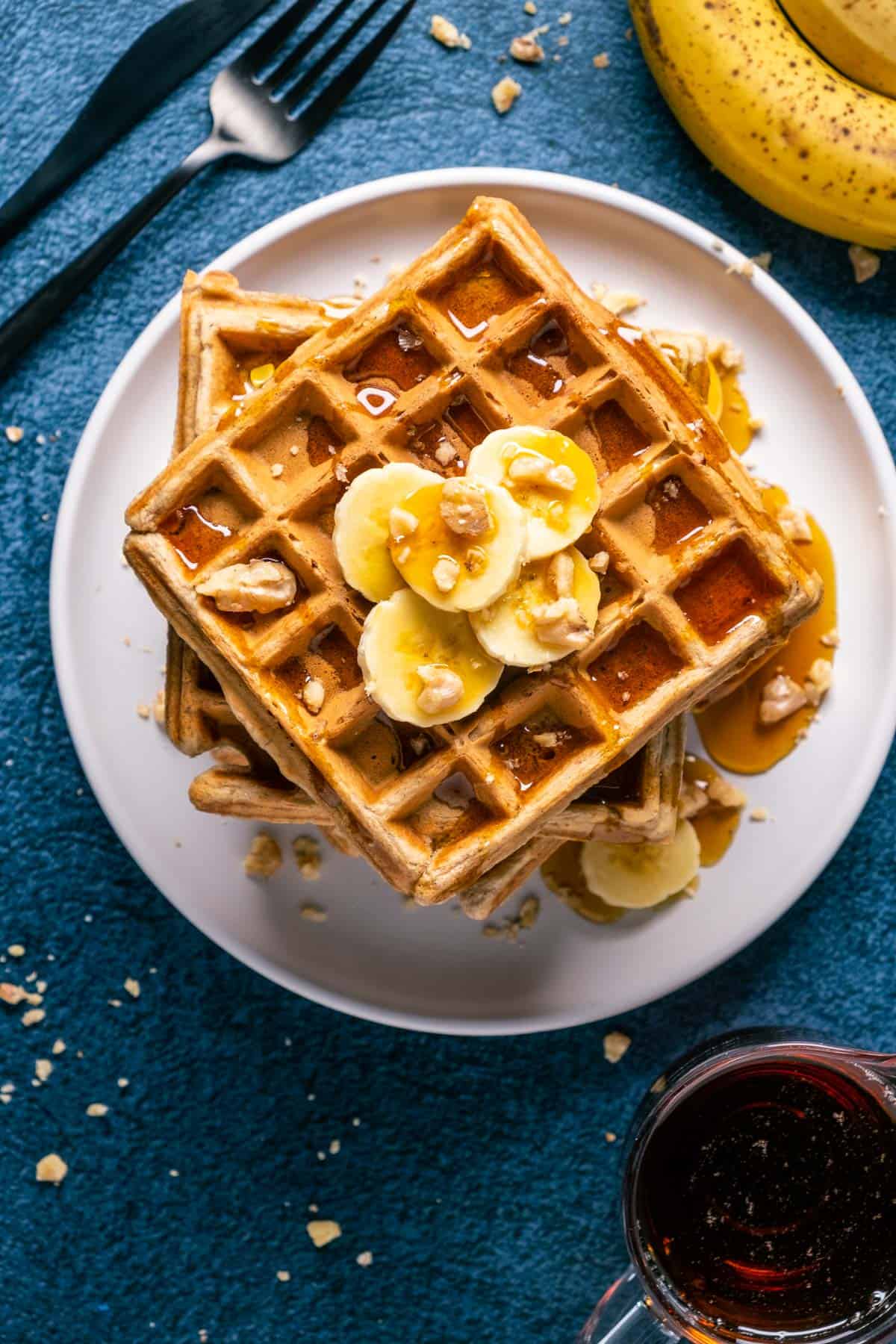 Overhead photo of a stack of vegan banana waffles topped with sliced banana, walnuts and syrup on a white plate.