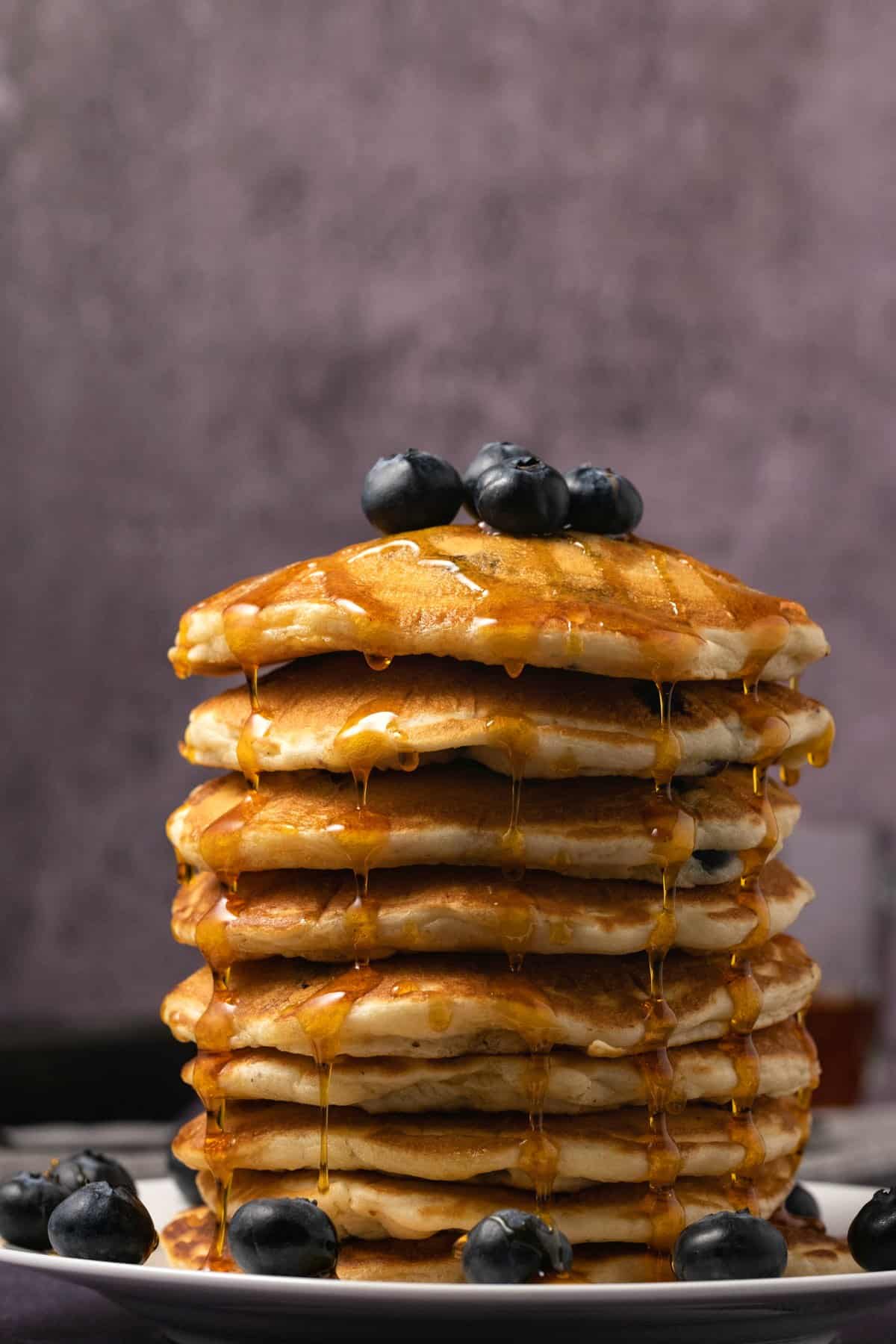 A stack of vegan blueberry pancakes drizzled with syrup and topped with fresh blueberries. 