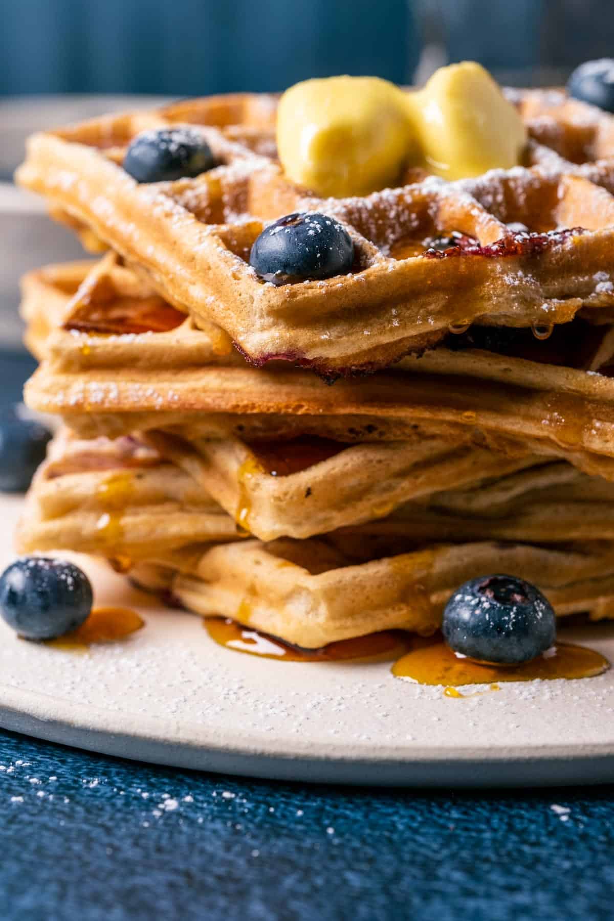 Stack of blueberry waffles topped with vegan butter, powdered sugar and syrup on a white plate.