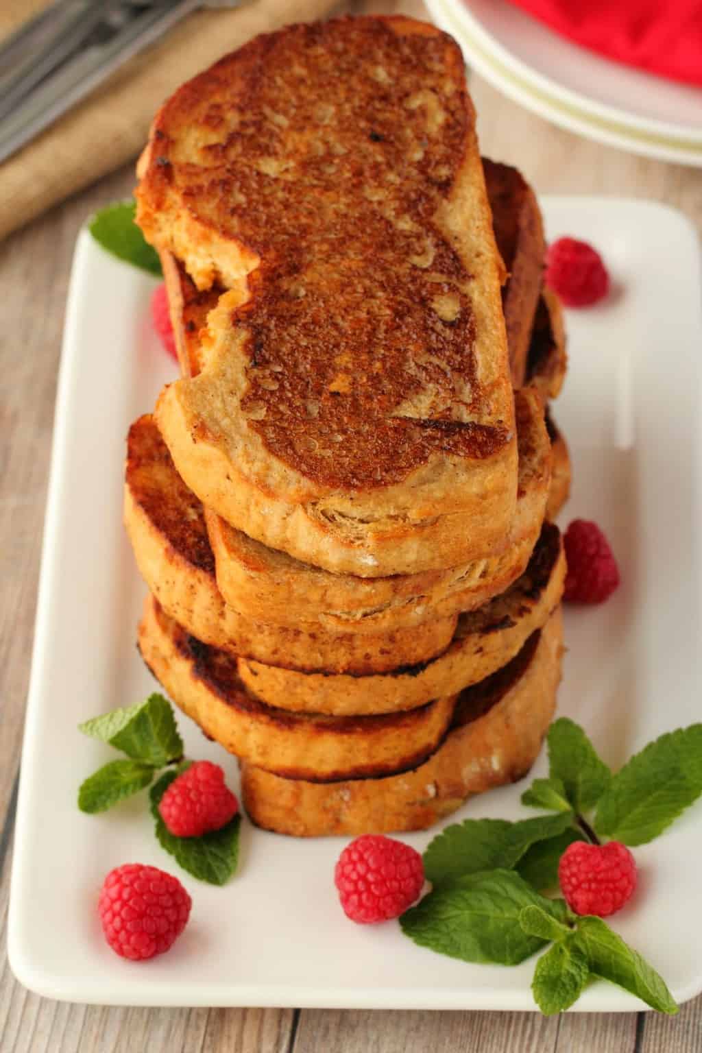 A stack of vegan french toast surrounded by mint leaves and raspberries, on a white plate. 
