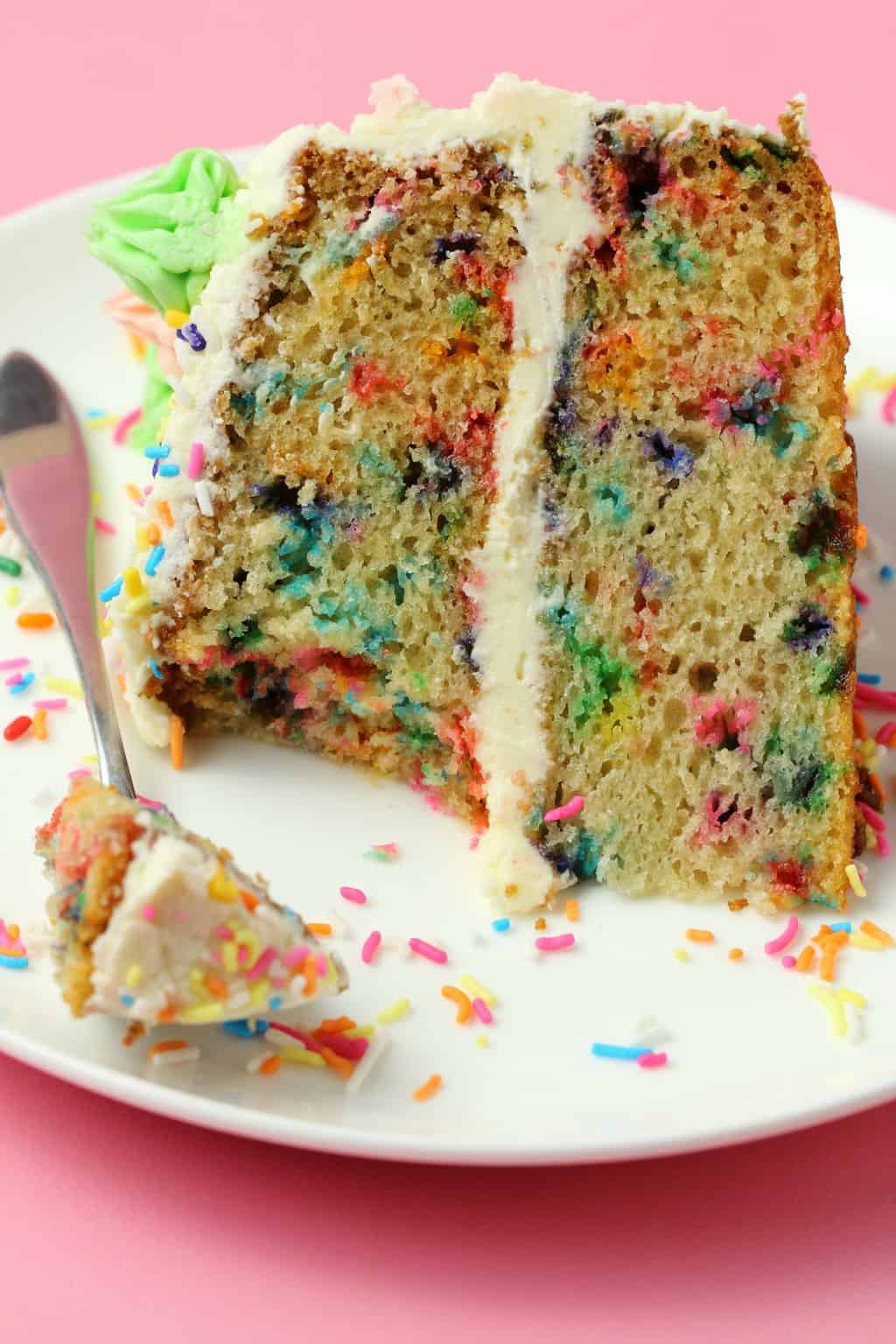 A slice of vegan funfetti cake on a white plate with a cake fork and sprinkles. 