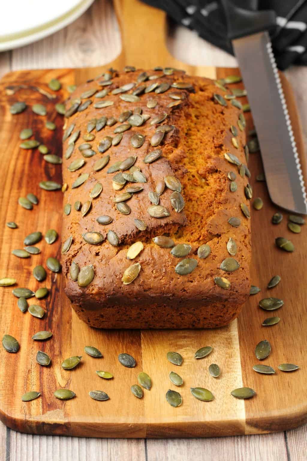 Vegan pumpkin bread topped with pumpkin seeds on a wooden board with a knife. 
