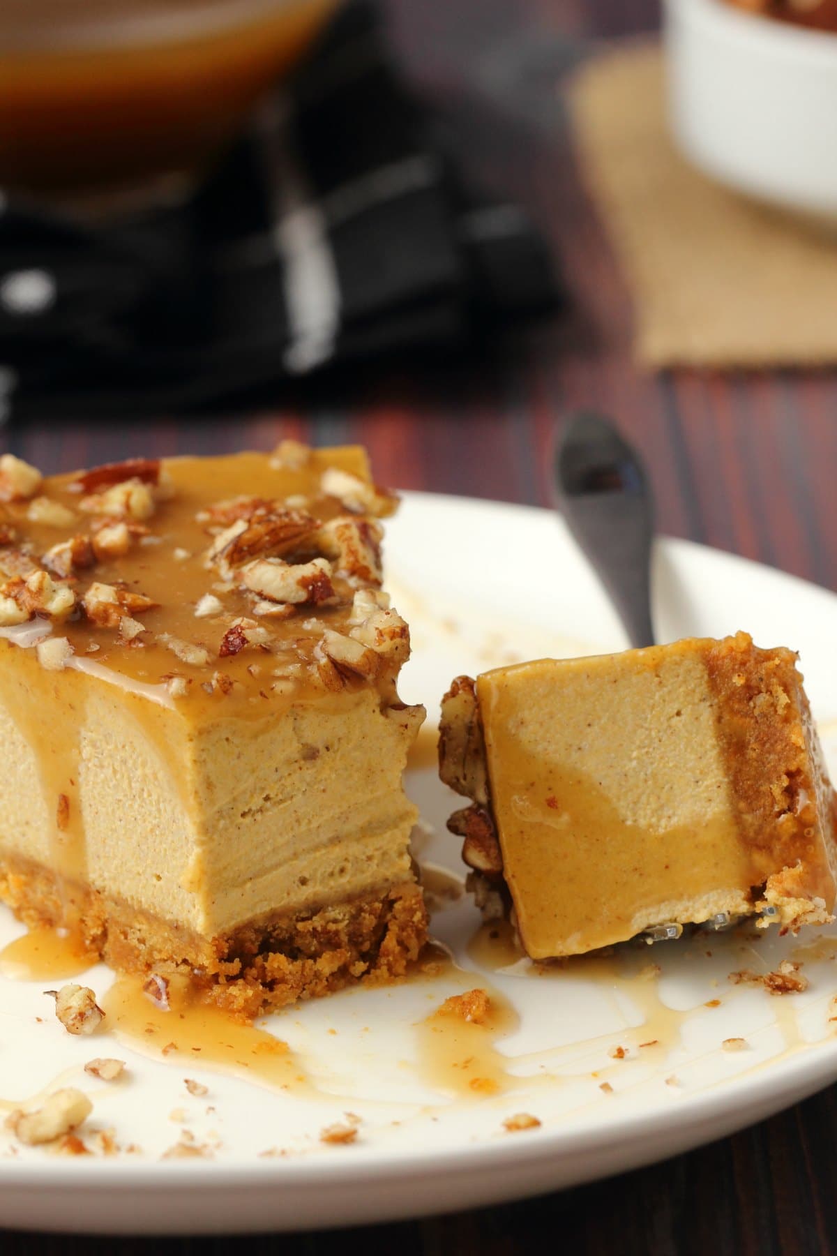 A slice of vegan pumpkin cheesecake topped with caramel sauce and crushed pecans on a white plate with a cake fork. 